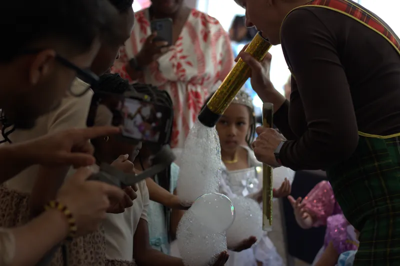 Children in party dresses watching a performer blow a tube of foam bubbles, with a gimbal and phone filming in the foreground